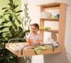 A baby is sitting on a wall-mounted changing table with shelves above it, next to a large plant. Produktbild-Vorschau