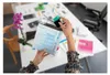 A person is holding an open envelope and inserting an ink cartridge into it, on a desk with various papers and a plant. Produktbild-Vorschau