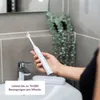 A person is holding a white electric toothbrush over a sink in a bathroom. Anteprima dell’immagine prodotto