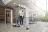 A woman is vacuuming a tiled floor in a modern building with a spiral staircase and large windows. Produktbild-Vorschau
