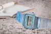 A light blue Casio digital watch is placed on a textured gray surface next to an open notebook and a pen. Náhled obrázku produktu