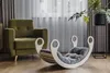A Montessori rocking chair with a grey cushion is placed on a wooden floor next to a green armchair, with sheer curtains in the background. Produktbild-Vorschau