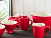 A collection of red ceramic kitchenware, including mugs and a mixing bowl, arranged on a dark surface. Produktbild-Vorschau