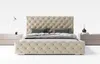 A tufted beige upholstered bed with two pillows and a duvet, placed between two nightstands with lamps and decorative items. Náhled obrázku produktu