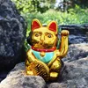 A golden Maneki Neko cat figurine sits on rocks outdoors, with greenery in the background. Produktbild-Vorschau