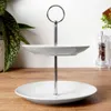 A two-tiered white porcelain cake stand with a silver handle is displayed on a wooden surface. A green plant is visible in the background to the left, and a salt and pepper grinder set is on the right. Podgląd zdjęcia produktu