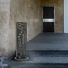 A decorative metal sign with the text "Herzlich Willkommen" (Welcome) is placed on the steps leading to a dark wooden door. Produktbild-Vorschau