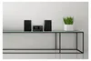 A compact audio system with two speakers and a central unit is displayed on a glass shelf against a white wall, with a potted plant to the right. Produktbild-Vorschau