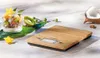 A kitchen scale with a bamboo top sits on a light-colored wooden surface next to a glass of fruit and yogurt and half a coconut. Náhľad obrázku produktu