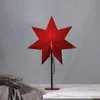 A red star-shaped lamp on a black stand, placed on a table with a grey cloth and dried branches. Podgląd zdjęcia produktu
