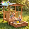 Two young children playing in a wooden sandbox with a roof and a small platform in a garden. Produktbild-Vorschau