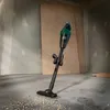 A cordless handheld vacuum cleaner is shown standing on a concrete floor in what appears to be a construction or renovation site. There are wood panels leaning against the wall in the background, and sawdust is scattered on the floor around the vacuum's no Produktbild-Vorschau