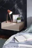 A bedside table with a lamp, picture frame, and plant, next to a bed with a book on it. Náhled obrázku produktu