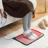A person is sitting on a couch, with their feet on a heated foot mat on a rug. Slippers are visible on the rug nearby. Náhled obrázku produktu