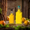 Two glass bottles filled with yellow liquid, surrounded by oranges, moss, and straw on a wooden surface. Produktbild-Vorschau
