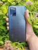 A hand holding a smartphone with a carbon fiber pattern on the back, surrounded by green leaves and pink flowers. Produktbild-Vorschau