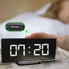 A person's hand is reaching to touch the top of a digital alarm clock displaying the time. Produktbild-Vorschau