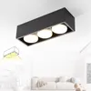 A modern black rectangular ceiling light with three circular white light fixtures, shown mounted on a white ceiling in a bright, minimalist living room setting with a sofa and a coffee table. Anteprima dell’immagine prodotto