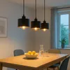 A dining table with a bowl of lemons, a carafe of water, and glasses, illuminated by three black pendant lights hanging above. Náhled obrázku produktu