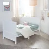 A white baby crib with bedding and pillows in a bright, airy room with artwork on the wall and a window with sheer curtains. Produktbild-Vorschau