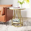 A round side table with a marble-like top and gold metal legs, placed next to a brown leather armchair. Books and a magazine are on the table, and a vase with greenery is visible in the background. Produktbild-Vorschau