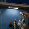 A hand holding a remote control points it towards LED lights under a kitchen cabinet. Bottles and utensils are visible on the counter below. Produktbild-Vorschau