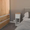 A narrow bedside table with a drawer and an open shelf sits next to a bed, with a wooden wardrobe on the left. Náhled obrázku produktu