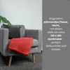 A red fleece blanket is draped over a gray armchair, with a dark gray pillow and a plant in the background. Náhled obrázku produktu