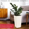 A cream-colored planter with a textured surface is shown in a living room setting, next to a sofa and a wooden cabinet. Podgląd zdjęcia produktu