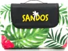 A folded picnic blanket with a tropical leaf and flower pattern, featuring a black carrying handle and flap with a logo. Náhled obrázku produktu