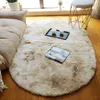 A fluffy oval rug with a gradient of beige and brown tones is placed on a wooden floor. A beige sofa is partially visible on the left, and a small dark tray with two white cups sits on the rug. A magazine or book is also on the rug. Náhled obrázku produktu