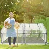 A woman is standing in a garden holding a basket of vegetables and a straw hat next to a greenhouse. Voir les images du produit