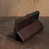 A dark brown leather phone case with a built-in stand is displayed on a textured surface. Anteprima dell’immagine prodotto