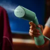 A hand holds a green garment steamer, emitting steam towards a maroon garment. Produktbild-Vorschau