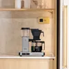 A MoccMaster coffee maker is shown on a kitchen counter, with shelves and decor in the background. Náhľad obrázku produktu