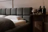 A close-up view of a dark gray upholstered bed with a white textured pillow and a small wooden side table with decorative bottles and bowls. Produktbild-Vorschau