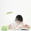 A young girl is studying at a desk with a dinosaur-shaped lamp. Produktbild-Vorschau