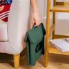 A hand holding a folded green stool next to a couch and a shelf. Produktbild-Vorschau