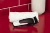 A hair clipper and a folded towel on a red countertop, with a tiled red wall in the background and a white ribbed container to the right. Podgląd zdjęcia produktu