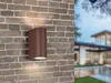 A brown outdoor wall light is mounted on a brick wall, with a blurred modern house and pool in the background. Voir les images du produit