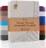 A folded fitted sheet is displayed prominently, with a band of text across the front. Several other fitted sheets in different colors are stacked behind it. Produktbild-Vorschau