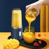 A hand is pouring orange juice from a portable blender cup into a glass, with a mango and strawberries on the side. Podgląd zdjęcia produktu