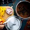 A close-up overhead view of a camping meal preparation scene, featuring a pot of food, sliced vegetables on a cutting board, and a pot lid. Produktbild-Vorschau