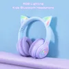A pair of purple and white cat-ear headphones with RGB lighting is displayed against a light blue and purple background. Podgląd zdjęcia produktu