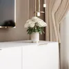 A white TV lowboard with a bouquet of white roses on top, placed in a modern living room setting with curtains and decorative lighting in the background. Náhled obrázku produktu