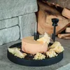 A cheese scraper tool is used to shave curls of cheese from a wheel of cheese, which sits on a black serving tray. The background shows a rustic setting with stacked stones and firewood. Produktbild-Vorschau