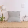 A large, light gray beanbag chair is placed on a light brown rug in front of a white wall. A floor lamp with a white shade is to the left, and a small side table with a decorative vase is to the right. A framed piece of art hangs on the wall. Voir les images du produit