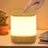 A hand is touching a wooden-topped LED lamp on a white surface, with a potted plant to the left and books to the right. Produktbild-Vorschau