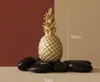 A golden pineapple ornament sits on dark stones against a two-toned background. Produktbild-Vorschau