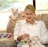 A woman is embroidering while a cat sits on a couch behind her. Produktbild-Vorschau